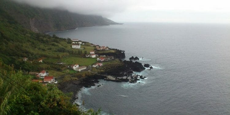 Un paesaggio costiero caratterizzato da piccole case bianche dai tetti rossi sparse sulle verdi scogliere dell'Ilha de São Jorge, a picco sul mare. Le onde dell'oceano si infrangono contro gli affioramenti rocciosi scuri, mentre la nebbia e le nuvole coprono parzialmente le montagne sullo sfondo.