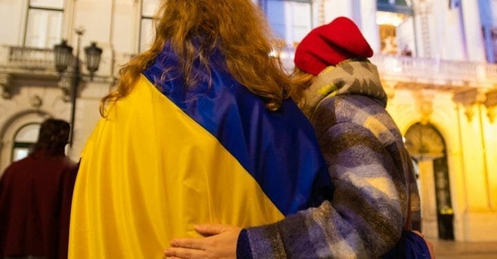 Due individui, visti da dietro, si abbracciano mentre stanno di fronte a un edificio. Uno è drappeggiato con una bandiera ucraina, che simboleggia la solidarietà in mezzo alla crisi e le opportunità per le offerte lavoro rifugiati ucraini, mentre l'altro indossa una giacca colorata e un berretto rosso. L'ambientazione sembra essere serale con l'edificio illuminato sullo sfondo.