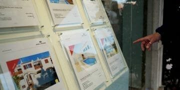 Una persona indica gli annunci immobiliari visualizzati in una finestra di vetro. Gli annunci presentano immagini di varie case e descrizioni dettagliate, comprese informazioni sul valore case portogallo. L'ambientazione sembra essere all'esterno, con il riflesso degli alberi nel vetro.