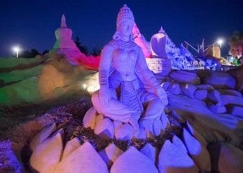 Di notte, una grande scultura di sabbia di una divinità seduta è illuminata da luci colorate, catturando il fascino di Attrazioni Algarve. La scultura è estremamente dettagliata, caratterizzata da gioielli e abiti decorati, con sculture circostanti e formazioni di sabbia visibili sullo sfondo.