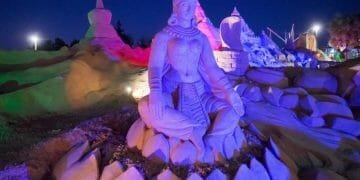 Di notte, una grande scultura di sabbia di una divinità seduta è illuminata da luci colorate, catturando il fascino di Attrazioni Algarve. La scultura è estremamente dettagliata, caratterizzata da gioielli e abiti decorati, con sculture circostanti e formazioni di sabbia visibili sullo sfondo.