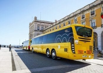 Un lungo autobus turistico giallo con il logo "Yellow Bus", della compagnia Trasporti Lisbona, è parcheggiato in un piazzale aperto accanto a un grande edificio storico con finestre ad arco e pilastri. Un altro autobus più piccolo della Carris Metropolitana è collegato nella parte posteriore, creando una formazione di autobus estesa.