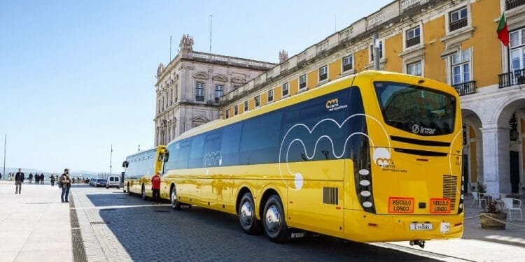 Un lungo autobus turistico giallo con il logo "Yellow Bus", della compagnia Trasporti Lisbona, è parcheggiato in un piazzale aperto accanto a un grande edificio storico con finestre ad arco e pilastri. Un altro autobus più piccolo della Carris Metropolitana è collegato nella parte posteriore, creando una formazione di autobus estesa.