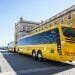 Un lungo autobus turistico giallo con il logo "Yellow Bus", della compagnia Trasporti Lisbona, è parcheggiato in un piazzale aperto accanto a un grande edificio storico con finestre ad arco e pilastri. Un altro autobus più piccolo della Carris Metropolitana è collegato nella parte posteriore, creando una formazione di autobus estesa.