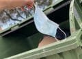 Una mano getta una maschera chirurgica azzurra in un cassonetto verde, aderendo alle misure di controllo della pandemia. Il cassonetto è parzialmente pieno di cartone e sacchi neri della spazzatura. Sullo sfondo sfocato sono visibili alberi e un cielo parzialmente nuvoloso.