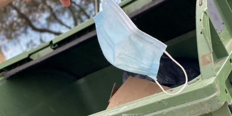 Una mano getta una maschera chirurgica azzurra in un cassonetto verde, aderendo alle misure di controllo della pandemia. Il cassonetto è parzialmente pieno di cartone e sacchi neri della spazzatura. Sullo sfondo sfocato sono visibili alberi e un cielo parzialmente nuvoloso.