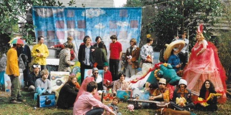 Un vivace gruppo di persone si riunisce all'aperto, vestito con una serie di costumi colorati e stravaganti, tra cui cappelli oversize, ali di fata e costumi di animali. Dietro di loro, un fondale dipinto raffigura nuvole e cielo. Vari oggetti di scena e decorazioni che ricordano IndieLisboa riempiono la scena, creando un'atmosfera festosa.