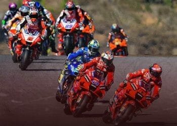 Diversi motociclisti in tute da corsa colorate e caschi percorrono una pista sulle loro moto ad alta velocità durante una gara competitiva del Moto GP Portogallo. I primi due corridori viaggiano su bici rosse, seguiti da vicino da altri su bici di vari colori. Lo sfondo è sfocato.