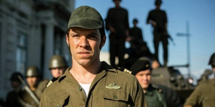 Un uomo in uniforme militare e berretto, somigliante a Salgueiro Maia, sta in primo piano con un'espressione seria. Dietro di lui sono visibili diversi soldati che indossano uniformi ed elmetti, alcuni in piedi su un veicolo. Lo sfondo mostra un edificio e il cielo è limpido e luminoso.