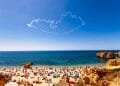 Una spiaggia affollata durante il picco del turismo in Algarve, con numerosi ombrelloni colorati e bagnanti sulla riva sabbiosa. Le onde blu dell'oceano si infrangono dolcemente sulla costa sullo sfondo, mentre una scia di fumo a forma di cuore adorna il cielo azzurro sopra.
