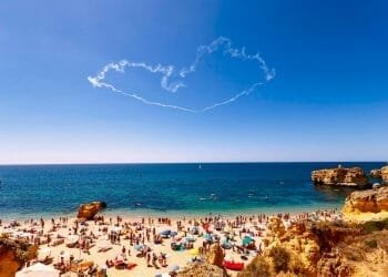 Una spiaggia affollata durante il picco del turismo in Algarve, con numerosi ombrelloni colorati e bagnanti sulla riva sabbiosa. Le onde blu dell'oceano si infrangono dolcemente sulla costa sullo sfondo, mentre una scia di fumo a forma di cuore adorna il cielo azzurro sopra.