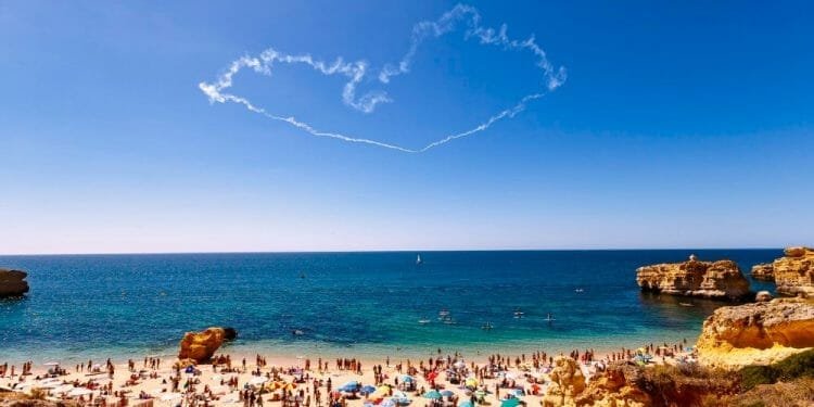 Una spiaggia affollata durante il picco del turismo in Algarve, con numerosi ombrelloni colorati e bagnanti sulla riva sabbiosa. Le onde blu dell'oceano si infrangono dolcemente sulla costa sullo sfondo, mentre una scia di fumo a forma di cuore adorna il cielo azzurro sopra.