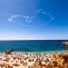Una spiaggia affollata durante il picco del turismo in Algarve, con numerosi ombrelloni colorati e bagnanti sulla riva sabbiosa. Le onde blu dell'oceano si infrangono dolcemente sulla costa sullo sfondo, mentre una scia di fumo a forma di cuore adorna il cielo azzurro sopra.