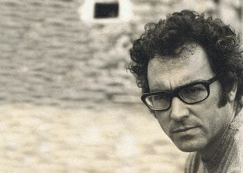 Un uomo con i capelli ricci e grandi occhiali dalla montatura scura guarda attentamente la telecamera. L'immagine, in tonalità seppia, emana un'atmosfera vintage. Lo sfondo è sfocato ma sembra essere un muro di pietra con una piccola finestra o apertura, che ricorda un'epoca in cui figure come José Afonso lasciarono il segno.