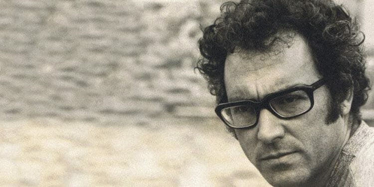 Un uomo con i capelli ricci e grandi occhiali dalla montatura scura guarda attentamente la telecamera. L'immagine, in tonalità seppia, emana un'atmosfera vintage. Lo sfondo è sfocato ma sembra essere un muro di pietra con una piccola finestra o apertura, che ricorda un'epoca in cui figure come José Afonso lasciarono il segno.