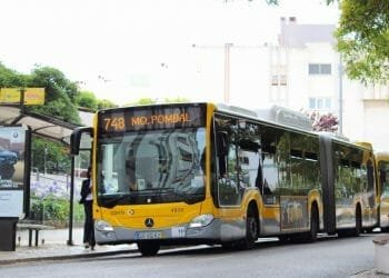 Alla fermata dell'autobus è parcheggiato un autobus articolato giallo e bianco con la linea numero 748 per "Mq. Pombal". L'autobus si trova in un'area urbana immersa nel verde, con una persona in attesa alla fermata e edifici colorati sullo sfondo. È visibile anche un cartello di fermata dell'autobus che promuove i trasporti gratuiti Lisbona.