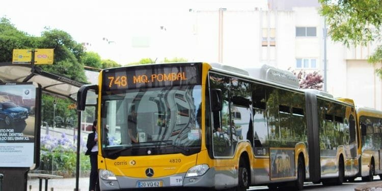 Alla fermata dell'autobus è parcheggiato un autobus articolato giallo e bianco con la linea numero 748 per "Mq. Pombal". L'autobus si trova in un'area urbana immersa nel verde, con una persona in attesa alla fermata e edifici colorati sullo sfondo. È visibile anche un cartello di fermata dell'autobus che promuove i trasporti gratuiti Lisbona.