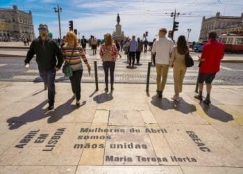 Persone che camminano su una grande piazza a Lisbona, Portogallo. In primo piano c'è un'iscrizione a terra che recita: "Abril em Lisboa Mulheres de Abril somos mãos unidas Maria Teresa Horta". Sullo sfondo sono visibili edifici, tram e una statua, tutti testimoni delle ultime regole e raccomandazioni.