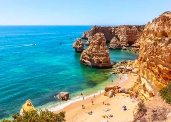 Una pittoresca spiaggia dell'Algarve con sabbia dorata, circondata da spettacolari scogliere e acque turchesi. Le persone prendono il sole e nuotano, mentre le barche navigano in lontananza. Il cielo limpido e le formazioni rocciose si aggiungono alla bellezza naturale di questo paesaggio balneare di Bandiere Blu.