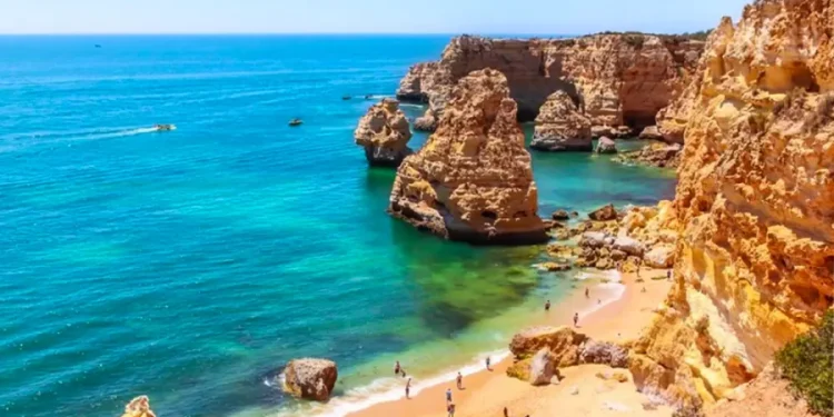Una pittoresca spiaggia dell'Algarve con sabbia dorata, circondata da spettacolari scogliere e acque turchesi. Le persone prendono il sole e nuotano, mentre le barche navigano in lontananza. Il cielo limpido e le formazioni rocciose si aggiungono alla bellezza naturale di questo paesaggio balneare di Bandiere Blu.