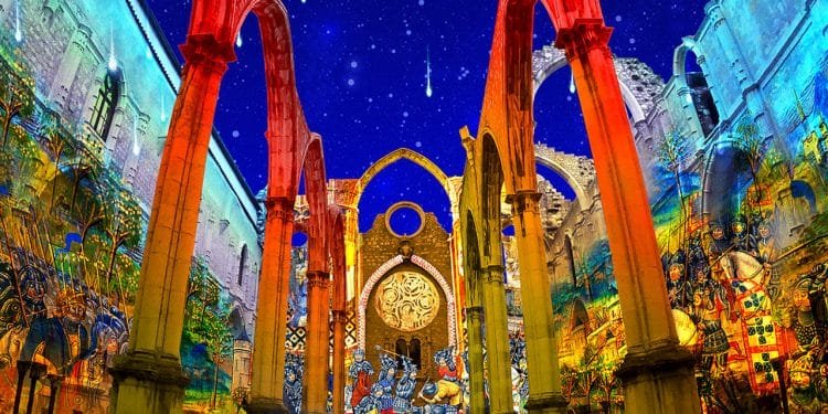 Un'immagine vibrante e notturna delle rovine di una chiesa storica con proiezioni colorate sulle pareti, raffiguranti varie scene e figure. Il pavimento è ricoperto di opere d'arte intricate, che ricordano Fado e spettacoli. In alto, il cielo stellato è ornato di stelle cadenti. Le rovine conservano alti archi e colonne.