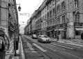 Foto in bianco e nero di una strada cittadina con vecchi edifici su entrambi i lati. I veicoli percorrono la strada e si vedono alcuni pedoni. In alto, lampioni e cavi decorativi attraversano la strada. L'atmosfera suggerisce una vecchia città europea, che ricorda vivere a Lisbona.