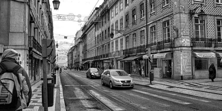 Foto in bianco e nero di una strada cittadina con vecchi edifici su entrambi i lati. I veicoli percorrono la strada e si vedono alcuni pedoni. In alto, lampioni e cavi decorativi attraversano la strada. L'atmosfera suggerisce una vecchia città europea, che ricorda vivere a Lisbona.