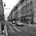 Foto in bianco e nero di una strada cittadina con vecchi edifici su entrambi i lati. I veicoli percorrono la strada e si vedono alcuni pedoni. In alto, lampioni e cavi decorativi attraversano la strada. L'atmosfera suggerisce una vecchia città europea, che ricorda vivere a Lisbona.