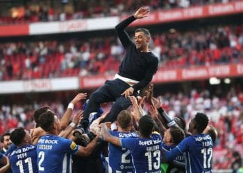 Una squadra di calcio vestita con maglie blu solleva il proprio manager, vestito con abiti scuri, in segno di festa. Lo sfondo presenta un grande stadio parzialmente pieno di tifosi. L'espressione dell'allenatore riflette la gioia mentre agita una mano, trionfante dopo aver vinto il campionato di calcio portoghese.