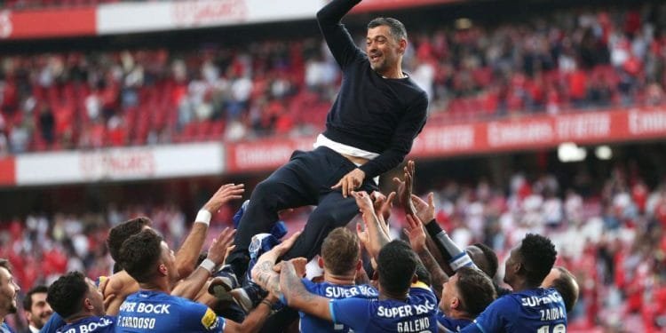 Una squadra di calcio vestita con maglie blu solleva il proprio manager, vestito con abiti scuri, in segno di festa. Lo sfondo presenta un grande stadio parzialmente pieno di tifosi. L'espressione dell'allenatore riflette la gioia mentre agita una mano, trionfante dopo aver vinto il campionato di calcio portoghese.