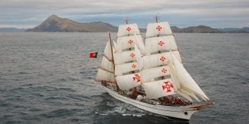 Un'alta nave con vele bianche con croci rosse, che sventola una bandiera rossa e verde, naviga in acque aperte durante la festa Marinha Portuguesa. Le montagne e il cielo coperto fanno da sfondo.