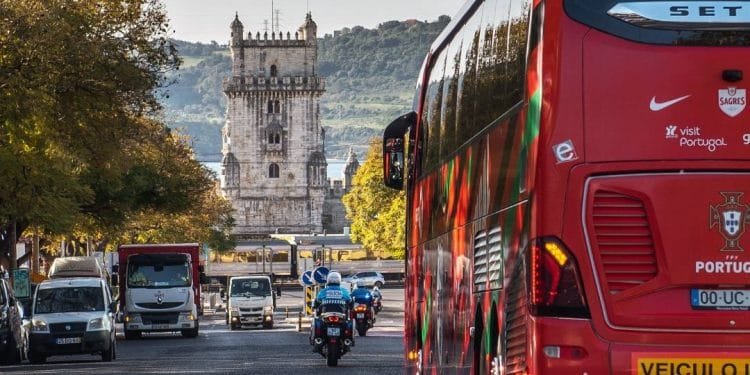 Un autobus rosso con la scritta "Portogallo" sul retro percorre una strada alberata a Lisbona. Un motociclista lo scorta e diversi veicoli procedono nella direzione opposta, rispettando i limiti di velocità. Sullo sfondo c'è una torre storica dall'architettura complessa.