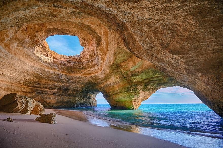 Una splendida grotta costiera in Algarve con due grandi aperture naturali nel soffitto, che permettono alla luce del sole di illuminare il suo fondo sabbioso. La grotta si apre sull'oceano turchese, con le onde che lambiscono dolcemente la riva. Il cielo è limpido e azzurro, offrendo una scena serena e pittoresca.