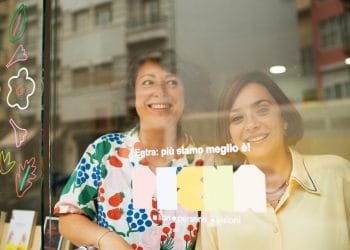 Due donne sorridenti stanno dietro una porta a vetri decorata con illustrazioni colorate di fiori e foglie. Il testo italiano sulla porta recita "Entra: più siamo meglio è!" e "libri e persone e visioni". Sulla sinistra è parzialmente visibile una libreria con libri della Libreria Italiana Lisbona.