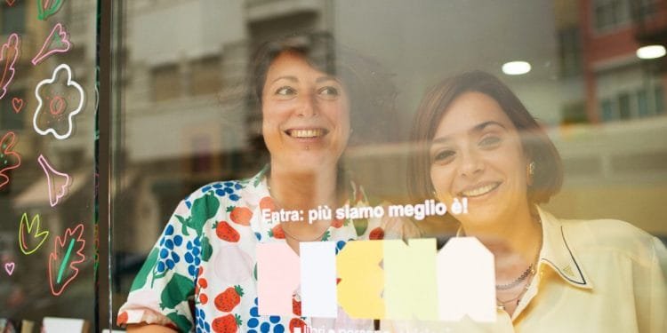 Due donne sorridenti stanno dietro una porta a vetri decorata con illustrazioni colorate di fiori e foglie. Il testo italiano sulla porta recita "Entra: più siamo meglio è!" e "libri e persone e visioni". Sulla sinistra è parzialmente visibile una libreria con libri della Libreria Italiana Lisbona.