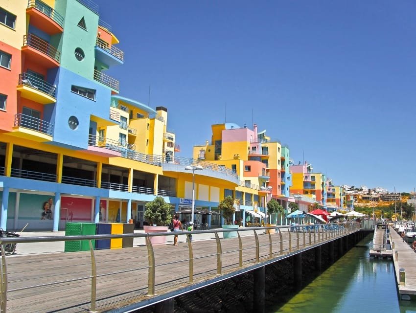 Una vivace scena sul lungomare presenta una fila di edifici moderni e colorati in blu, giallo, rosa e verde. Una passerella di legno con ringhiera di metallo corre lungo l'acqua, con persone che passeggiano e negozi e caffè visibili. Perfetto per esplorare le Attrazioni Algarve, il cielo è limpido e azzurro.
