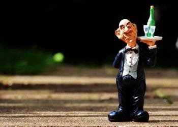 Una piccola statuetta di un cameriere vestito con uno smoking nero con un papillon, che tiene in mano un vassoio con una bottiglia verde e due bicchieri. La statuina, che ricorda la ristorazione in Portogallo, ha lineamenti del viso esagerati e si trova su una superficie strutturata con la luce del sole che proietta ombre sullo sfondo.