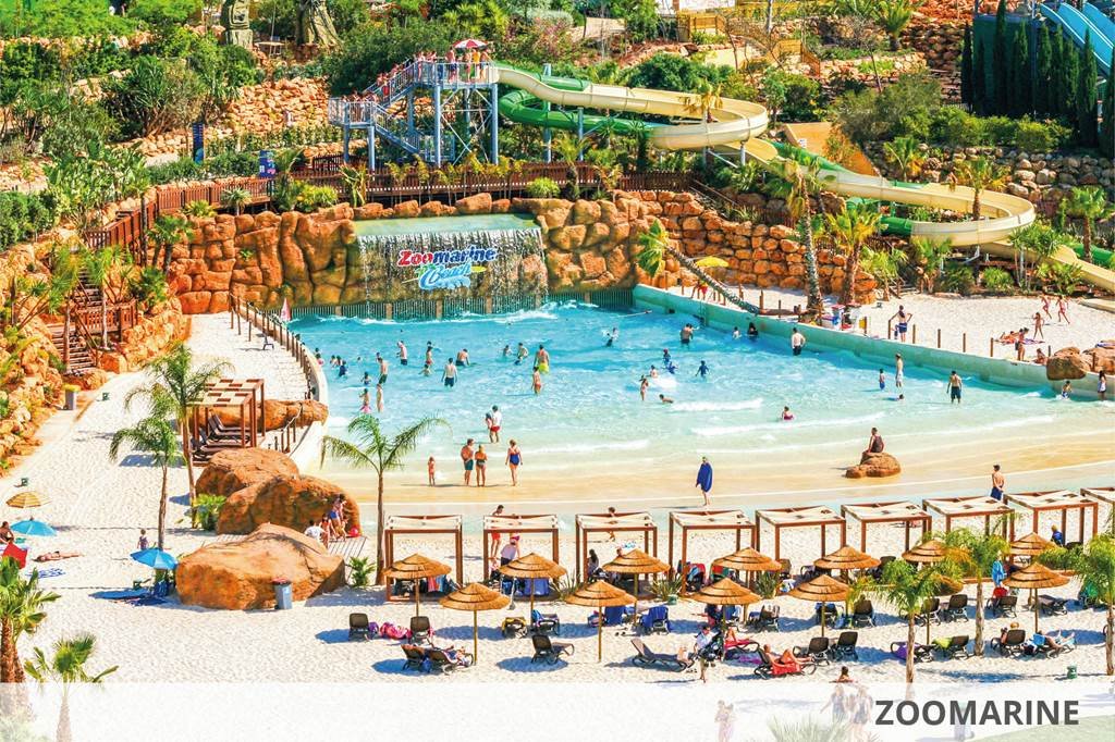 L'immagine raffigura una vivace scena del parco acquatico di Zoomarine, una delle principali attrazioni che l'Algarve ha da offrire. È dotato di una grande piscina a onde circondata da persone che si godono l'acqua. Uno scivolo acquatico tortuoso, una vegetazione lussureggiante e ombrelloni di paglia con sedie a sdraio riempiono lo sfondo. L'ambiente è soleggiato e vivace.