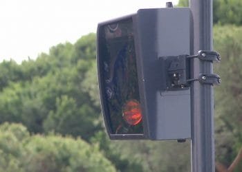 Un primo piano di una telecamera autovelox montata su un palo a Lisbona, con una zona boschiva visibile sullo sfondo. La fotocamera è nera e presenta un copriobiettivo rettangolare, con una piccola luce rossa illuminata all'interno.