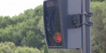 Un primo piano di una telecamera autovelox montata su un palo a Lisbona, con una zona boschiva visibile sullo sfondo. La fotocamera è nera e presenta un copriobiettivo rettangolare, con una piccola luce rossa illuminata all'interno.