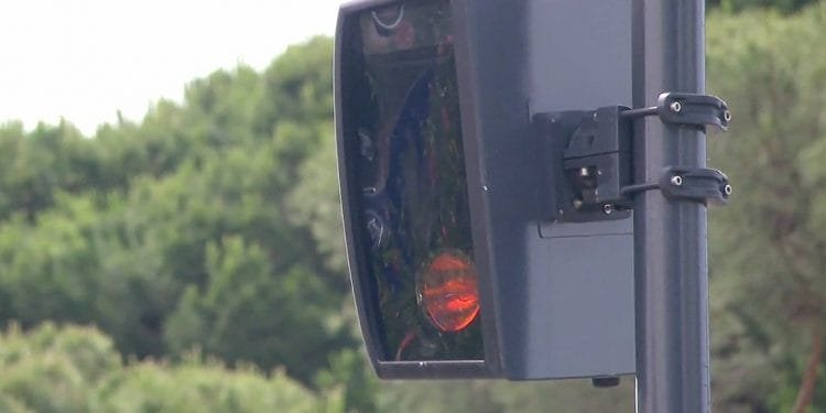 Un primo piano di una telecamera autovelox montata su un palo a Lisbona, con una zona boschiva visibile sullo sfondo. La fotocamera è nera e presenta un copriobiettivo rettangolare, con una piccola luce rossa illuminata all'interno.