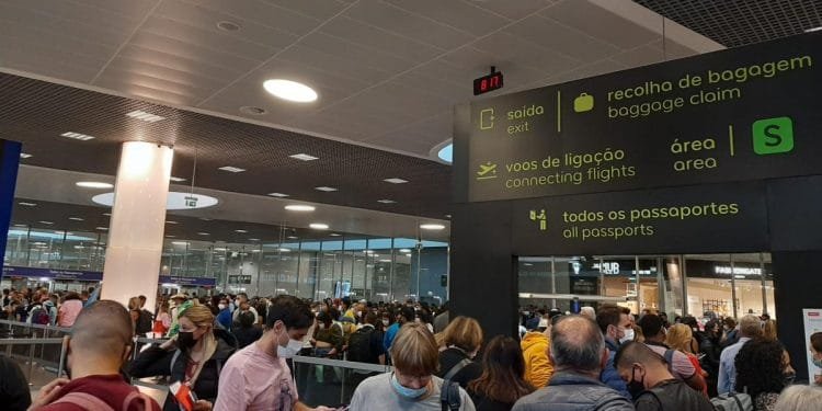 Un terminal aeroportuale affollato con viaggiatori in fila. I segnali indicano le indicazioni per le uscite, i voli in coincidenza, il ritiro bagagli e tutti i passaporti. Le persone indossano maschere, alcune hanno con sé documenti di viaggio. Un orologio digitale mostra l'ora mentre un annuncio menziona un problema all'aeroporto di Lisbona.