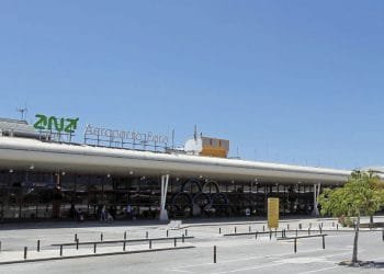 Immagine dell'esterno dell'aeroporto di Faro. L'edificio presenta un grande cartello con la scritta "Aeroporto de Faro" insieme a un logo verde. Il cielo è limpido e azzurro e il traffico veicolare e pedonale davanti al terminal è minimo. Questo vivace hub è anche conosciuto come Aeroporto Gago Coutinho.