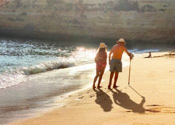 Una coppia di anziani, con indosso un cappello, cammina mano nella mano lungo una spiaggia sabbiosa con dolci onde che lambiscono la riva. Uno di loro usa un bastone da passeggio. Il sole è basso, proietta lunghe ombre e illumina le scogliere sullo sfondo: una scena serena per i pensionati esteri che si godono i loro anni d'oro all'estero.