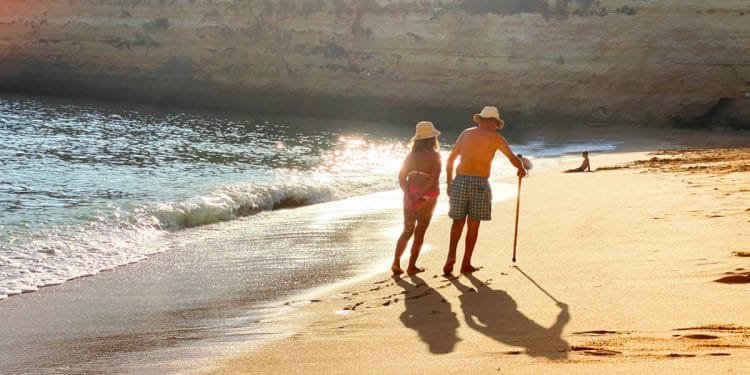 Una coppia di anziani, con indosso un cappello, cammina mano nella mano lungo una spiaggia sabbiosa con dolci onde che lambiscono la riva. Uno di loro usa un bastone da passeggio. Il sole è basso, proietta lunghe ombre e illumina le scogliere sullo sfondo: una scena serena per i pensionati esteri che si godono i loro anni d'oro all'estero.