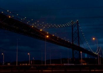 Un grande ponte sospeso è illuminato da luci gialle contro un cielo notturno scuro e nuvoloso. Il ponte si estende sull'immagine dall'angolo in alto a sinistra all'angolo in basso a destra. In basso sono visibili i lampioni e una sagoma del paesaggio, il tutto splendente sotto un piano di risparmio energetico.