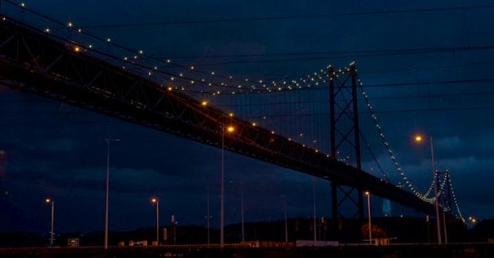 Un grande ponte sospeso è illuminato da luci gialle contro un cielo notturno scuro e nuvoloso. Il ponte si estende sull'immagine dall'angolo in alto a sinistra all'angolo in basso a destra. In basso sono visibili i lampioni e una sagoma del paesaggio, il tutto splendente sotto un piano di risparmio energetico.