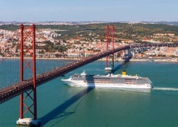 Una grande nave da crociera bianca, premiata dai World Travel Awards, naviga sotto un ponte sospeso rosso che attraversa un ampio fiume. Lo sfondo mostra un paesaggio urbano con numerosi edifici e colline verdi. Il cielo è limpido, mettendo in risalto la bellezza paesaggistica della migliore destinazione turistica del Portogallo.