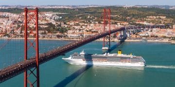 Una grande nave da crociera bianca, premiata dai World Travel Awards, naviga sotto un ponte sospeso rosso che attraversa un ampio fiume. Lo sfondo mostra un paesaggio urbano con numerosi edifici e colline verdi. Il cielo è limpido, mettendo in risalto la bellezza paesaggistica della migliore destinazione turistica del Portogallo.