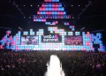 La grande folla di fronte al palco del Forum di Parque das Nações il durante il Web Summit di Lisbona 2024. Lo sfondo è caratterizzato da pannelli luminosi rettangolari colorati in tonalità blu, rosa e viola. Due grandi schermi mostrano le immagini dell'evento.